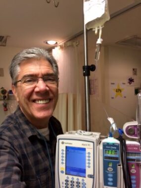 A man with salt-and-pepper hair and wearing glasses and a gray-and-black plaid shirt stands smiling beside an IV pole in what appears to be a hospital.