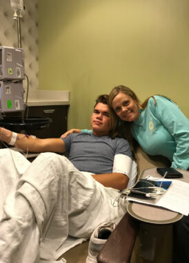 A different 17-year-old (the twin brother of the person pictured in the previous photo) reclines in an infusion chair at the clinic. The lower half of his body is covered with a blanket to keep him warm, and the therapy is delivered via drip infusion into his arm. His mom is pictured to his left.