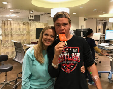 A 17-year-old with a backward baseball cap eats an orange popsicle in an infusion treatment center at a hospital. Next to him on his right is his mom. He has a black T-shirt that says "Outlaw." 