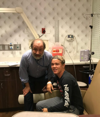 The same teenager that is pictured in the first photo above is seen here sitting down in an infusion chair and posing for the photo with an older doctor who is balding and has a gray beard. Both men are dressed casually.