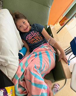 A young, brown-haired girl lies in a green lounge chair, with an orange blanket with green swirling stripes over her hips and legs. Her left arm is hooked up to an IV. A pillow and what may be a tumbler is on her right. She is smiling and wears a black T-shirt with pink writing, the first word of which is "Girls." A sandal with pink straps is on the yellowish floor in front of the chair.