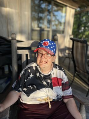 A man sits on a gray wooden chair on what appears to be a porch; part of the structure, with a large window, is behind him. He wears maroon pants, a T-shirt with parts of the U.S. flag, and a cap with white stars in a blue field, a red bill, and a red smiley face in the center.