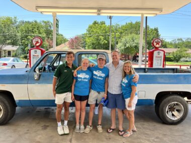 A group of five, all in shorts, stand in front of a blue and white, extra-large truck, all in front of a couple of old gas pumps.