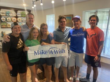 Three people of a group of seven hold a a white Make-A-Wish banner with blue lettering. They appear to be in a home, and those in the front row all are wearing shorts and T-shirts (though with one woman, much of her clothing is behind the banner). Two of the people appear to be adults.