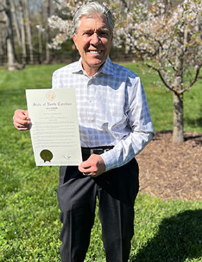 A man stands outside holding up an official proclamation from North Carolina's governor, stating that April is Fabry Disease Awareness Month. The man is wearing a checkered, collared shirt and dark slacks and is smiling proudly at the camera.