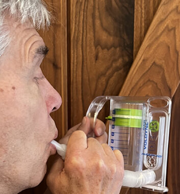 A close-up side view of a man with gray hair blowing into a spirometer device, which looks like a type of measuring cup with a white, plastic tube sticking out of it. The man holds the mouthpiece of the tube in his mouth and has his lips puckered, as if he's blowing into the tube. 