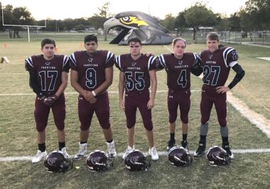 Five young men, all in high school football uniforms, stand in a horizontal line on a football field. Their helmets are on the ground in front of their feet. A large inflatable falcon head is on the field in the background, with a tunnel through the middle where the players can run.