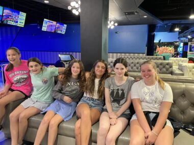 Six girls, ages 13 or 14, sit side by side on a brown couch in a bowling alley. Several screens displaying scores are visible in the background, and the alley is lit up in a deep blue. The girls are all smiling and have their arms around each other.