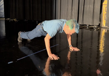 A man with blue and green hair and a matching blue T-shirt does a pushup on a stage. 