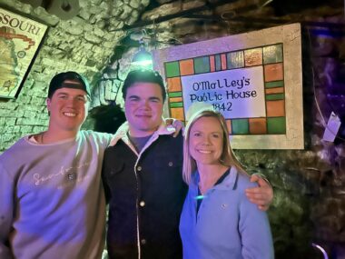 Three people — two men and a woman — stand in front of a room's sign that reads "O'Malley's Public House 1842." The roof and walls are stones, which look green because of the lighting; the roof is angled. They wear, from the men on the left, a gray-appearing sweatshirt and a ball cap worn backwards; a dark-colored jacket with a large white collar and what appears to be a blue T-shirt underneath; and a light blue jacket.