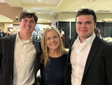 Two young men in suit jackets and white dress shirts flank a woman with blond hair and wearing a dark top. They're in some sort of public place.