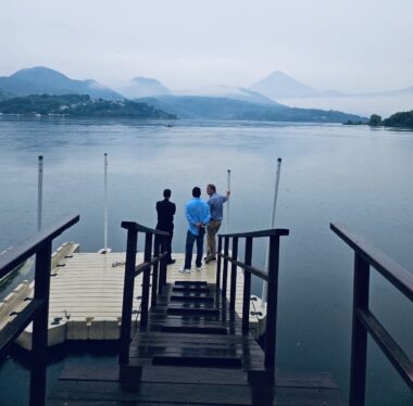 Three men stand on a dock looking out over a vast blue lake, with mountains in the distance. It is a majestic scene.