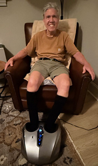 An older man with white-gray hair sits in an armchair with his feet in an electric foot massager. He's wearing a T-shirt and shorts and is smiling at the camera.