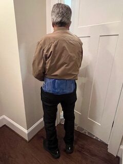 A man in a brown jacket stands with his back to the camera. He's wearing a blue ice pack around his lower back.
