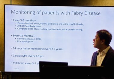 A phone photo of a man delivering a presentation on a large digital screen. He's looking back over his right shoulder at the screen while standing at a podium.