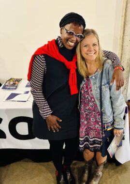 Two women, smiling broadly, embrace in a side hug in front of a table covered with promotional materials. 