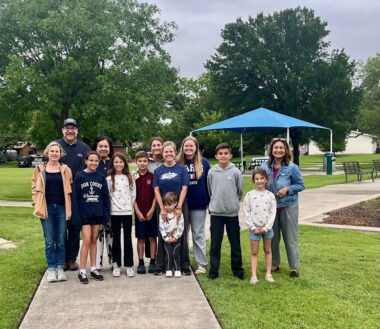 A group of people, both adults and children, pose for a photo on a sidewalk in a park.