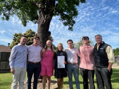 A family of seven — mother, father, four sons, and one daughter — stand side by side with their arms around each other. The mom is in the middle and holds up a document proclaiming April to be Fabry Awareness Month in Texas. The family is standing outside under a large tree, and it looks to be a beautiful day with blue skies.