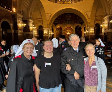Three women and one man happily pose for a photo in an ancient cathedral. They stand left to right with their arms interlocked. One of the women and the man are dressed in clothing used by members of the Order of Malta. 